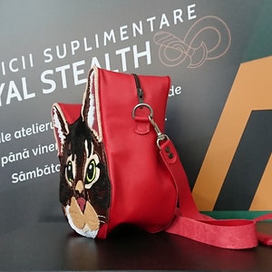 May include: A red leather handbag with a cat face design. The cat has brown and white fur and green eyes. The bag has a long red strap.