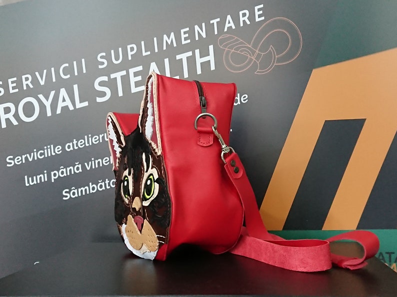 May include: A red leather handbag with a cat face design. The cat has brown and white fur and green eyes. The bag has a long red strap.