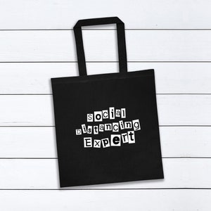 May include: Black tote bag with white text that reads "social distancing expert" in a ransom note style font.
