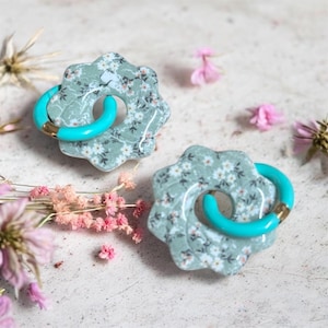 May include: A pair of distinctive earrings. Each earring has a floral pattern in green and white, with a bright turquoise hoop and gold-coloured accents. The earrings are set against a neutral background with dried flowers.
