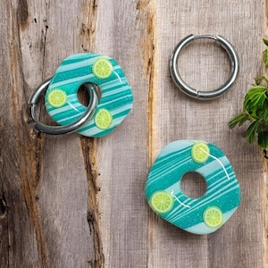 May include: A pair of earrings with a lime slice design. The earrings are a light blue and green color with silver hoops. The earrings are on a wooden surface.