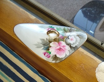 May include: A white porcelain dish with a gold rim and floral design. The dish is shaped like a triangle and has a small gold ring in the center. The flowers are pink, white, and green.