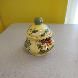 May include: A yellow ceramic sugar bowl with a green lid and floral design. The bowl has a scalloped edge and is decorated with green leaves, brown flowers, and yellow accents.