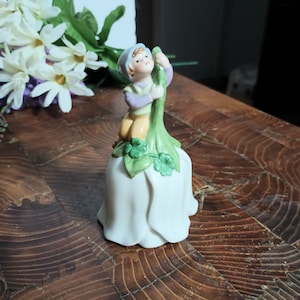 May include: A porcelain figurine of a boy in a blue hat sitting on a white bell with green leaves and a green stem. The bell is decorated with shamrocks.
