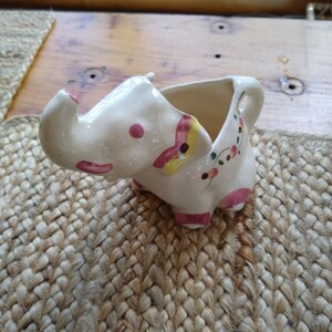 May include: A white ceramic elephant pitcher with pink, yellow, and green floral designs. The pitcher has a spout and a handle.