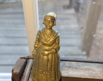 May include: A brass figurine shaped like a person wearing a bonnet, a shawl, and a long skirt with a decorative band near the hem. The metallic surface has a golden tone with visible signs of aging and patina, showing a standing figure.