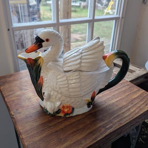 May include: A white ceramic swan teapot with a black and orange beak, and green and orange accents. The teapot has a detailed wing design and a small swan figurine on the base. The handle is green and black.