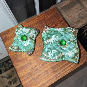 May include: Two square ceramic dishes with a turquoise and brown mottled glaze. Each dish has a large, round, green glass bead in the center. The dishes are displayed on a wooden surface.