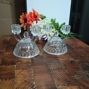 May include: Two clear glass candle holders with a diamond pattern base and three candle cups. The candle holders are on a wooden surface with colorful flowers in the background. The candle holders have a curved design.