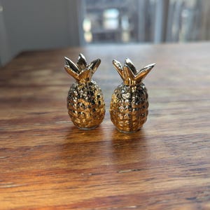 May include: Two small, gold-colored pineapple figurines with textured surfaces and detailed leafy crowns. The decorative pineapples are displayed on a wooden table, capturing a tropical theme.