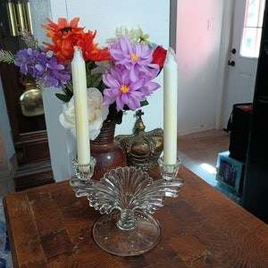 May include: A clear glass candle holder with two white candles and a decorative floral arrangement with orange, purple, and white flowers.