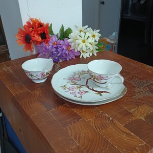 May include: A vintage teacup and saucer set with a floral design. The white porcelain cup and saucer feature a delicate pink cherry blossom branch and gold trim. A matching teacup is also present. Artificial flowers in orange, purple, and white are in the background.