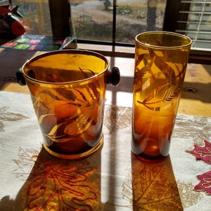 May include: Two amber glass containers with gold leaf designs. The container on the left is a bucket with a handle and the container on the right is a tall glass. Both containers are sitting on a white tablecloth with a brown and gold leaf pattern.