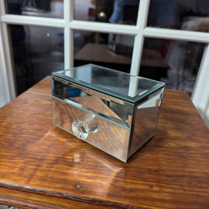May include: A small mirrored jewelry box with a clear crystal knob. The box has a drawer and a mirrored top.
