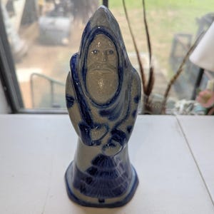 May include: A ceramic figurine of a bearded figure wearing a hooded cloak, featuring a light gray base with dark blue painted details. The figure has a pointed hood, a long beard, and a stylized tree design painted in blue at the bottom base.