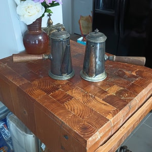 May include: Two antique silver coffee pots with wooden handles on a wooden cutting board.