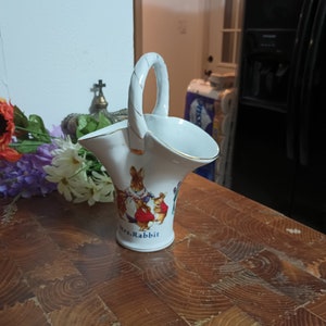 May include: White ceramic vase with a gold handle and a depiction of Mrs. Rabbit and her children from Beatrix Potter's "The Tale of Peter Rabbit".