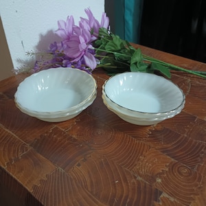 May include: Two white milk glass bowls with a gold rim. The bowls are on a wooden surface.
