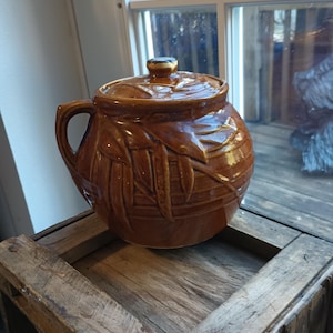 May include: A brown ceramic cookie jar with a lid and a leaf design. The jar is sitting on a wooden crate.