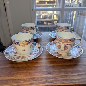 May include: Four matching teacups and saucers with floral and geometric patterns. The cups have gold handles and a blue and gold trim. The saucers have a matching design with a blue and gold border. The set is on a wooden surface.