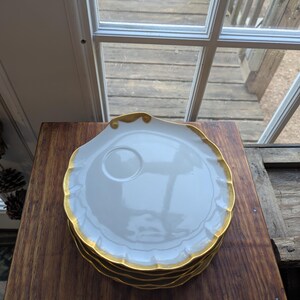 May include: A stack of white porcelain plates with scalloped edges and gold trim. Each plate has a small, circular indentation in the center. The plates are stacked on a dark wooden surface.