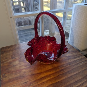 May include: A ruby red glass basket with a twisted handle. The basket has a ruffled edge and sits on a wooden surface. The glass has a textured appearance, and the handle is arched.