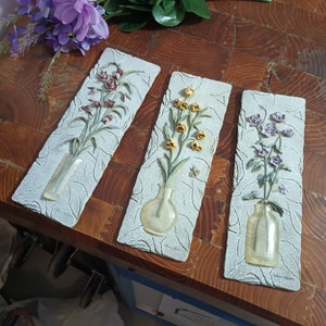 May include: Three decorative wall plaques with floral designs. Each plaque features a vase with a different type of flower, including purple, yellow, and pink flowers. The vases are made of a clear glass-like material and have a textured surface. The plaques are made of a light gray material with a textured surface.