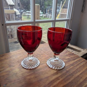 May include: Two ruby red wine glasses with a textured, beaded base and a rounded bowl. The glasses are set on a wooden surface, with a window and outdoor scenery in the background.