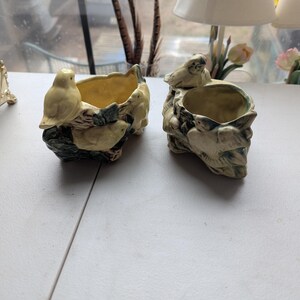 May include: Two ceramic bird planters. One planter features three yellow birds perched on a branch with green leaves. The other planter has a bird on the side and a textured, tree-like design. Both have a yellow interior.