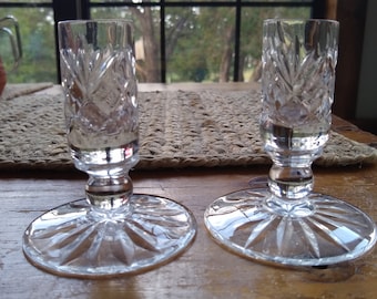 May include: Two clear glass candle holders with a cut glass design. The holders have a round base and a tall, cylindrical body.