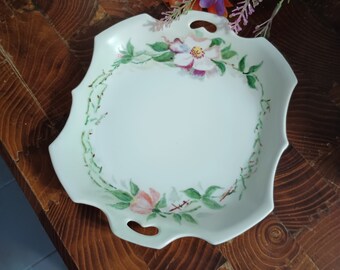 May include: A white porcelain serving dish with a scalloped edge and two handles. The dish is decorated with a hand-painted floral design featuring pink and white flowers and green leaves.