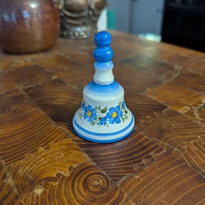 May include: A small, blue and white ceramic bell with a floral design. The bell has a blue knob on top and is decorated with blue flowers on a white background.