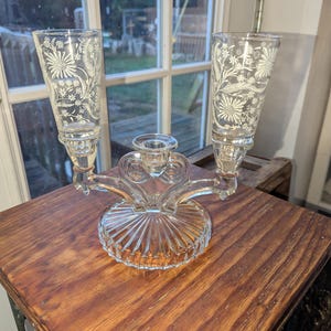 May include: A clear glass candelabra with two tall, flared candle holders and a central candle cup. The candle holders feature a white floral design. The base is round with a ribbed pattern.