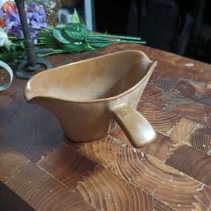 May include: A brown ceramic gravy boat with a handle. The boat is on a wooden surface.