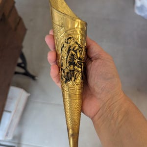 May include: A conical brass wall pocket featuring a textured surface and a central embossed panel depicting figures in a scene. The metal object has a pointed base and an open top, with intricate detailing throughout the gold-toned exterior.
