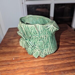 May include: Green ceramic planter shaped like a tree stump. The planter has a textured surface with carved details, including the word "STMW" and a heart. The planter is designed to hold plants or flowers.