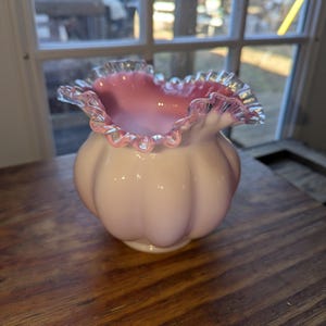 May include: A vintage, light pink glass vase with a ruffled rim. The vase has a bulbous body with a scalloped design. The rim is a darker pink with a clear, textured edge. The vase sits on a wooden surface.