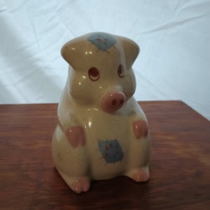 May include: A ceramic piggy bank with a light brown glaze and blue and red painted patches. The pig has pink hooves and a pink snout.