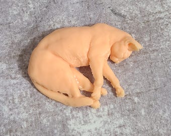 May include: A peach-coloured cat figurine in a curled-up sleeping position. The cat's paws are crossed, and its head is resting on its front paws. The figurine is smooth and appears to be made of a solid material.
