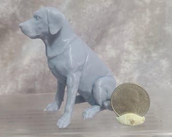 May include: A light grey dog figurine in a seated position. The dog is a Labrador or similar breed, with detailed features. A U.S. quarter coin is next to the dog for size comparison. The figurine is on a clear surface.