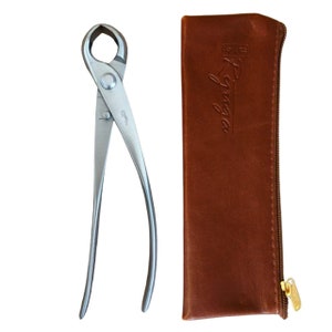 以下が含まれることがあります： ステンレス製の剪定ばさみと、茶色の革製の収納ケースのセット。ケースはジッパーで閉じることができ、前面には「Sassafras Co.」という文字が刻印されています。