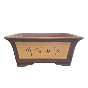 Puede incluir: Una maceta rectangular para bonsái con exterior marrón y panel central beige. El panel central presenta caligrafía negra. La maceta tiene un diseño ligeramente acampanado y se apoya en cuatro pequeños pies.