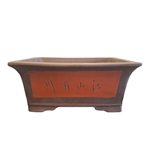 以下が含まれることがあります： 濃い茶色の外観と赤茶色の中心パネルを備えた長方形の盆栽鉢。 パネルには日本の書道が施されています。 鉢の内側は薄茶色で、小さな足の上に置かれています。