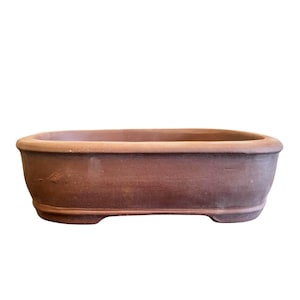 Może przedstawiać: Owalna doniczka do bonsai wykonana z terakoty o matowym, ziemistobrązowym wykończeniu. Pojemnik posiada szeroki, płaski brzeg, lekko zwężające się boki oraz podwyższoną podstawę z małymi nóżkami, zapewniając stabilne podparcie dla roślin.