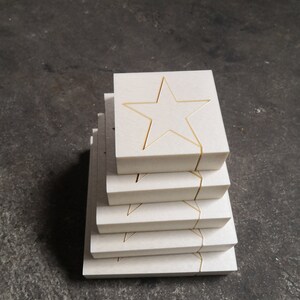 May include: A stack of six white square tiles with a gold star cutout in the center of each tile.