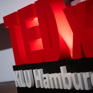 Könnte beinhalten: Große, rote "TEDx" Buchstaben über dem weißen Text "KLU Hamburg". Die Buchstaben sind beleuchtet und erzeugen einen auffälligen visuellen Effekt. Der Hintergrund ist neutral weiß und schwarz.