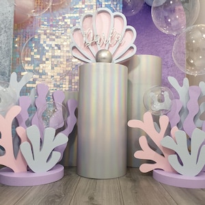 Può includere: Un espositore decorativo con un grande piedistallo cilindrico iridescente sormontato da una sagoma a forma di conchiglia rosa chiaro e bianca. La conchiglia riporta il nome Darla in corsivo bianco. Attorno al piedistallo ci sono sagome di corallo rosa chiaro e blu.