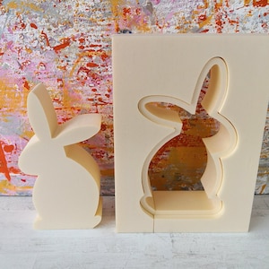 May include: Two white foam bunny shapes, one solid and one with a cutout. The solid bunny is on the left and the cutout bunny is on the right. Both bunnies are facing the right side of the image.