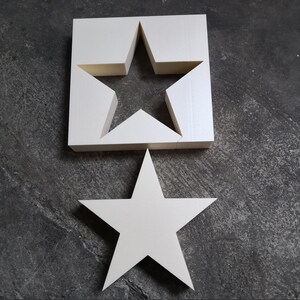 May include: Two white wooden stars, one with a cutout star shape. The larger star is on top of the smaller star.