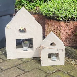 Peut inclure: Deux bougeoirs en béton blanc en forme de maison avec de petits bougeoirs en verre à l'intérieur. La plus grande maison a une petite fenêtre et une ouverture ronde au sommet. La plus petite maison a une ouverture ronde au sommet et une fenêtre rectangulaire. Les deux maisons ont un détail en ficelle rustique.
